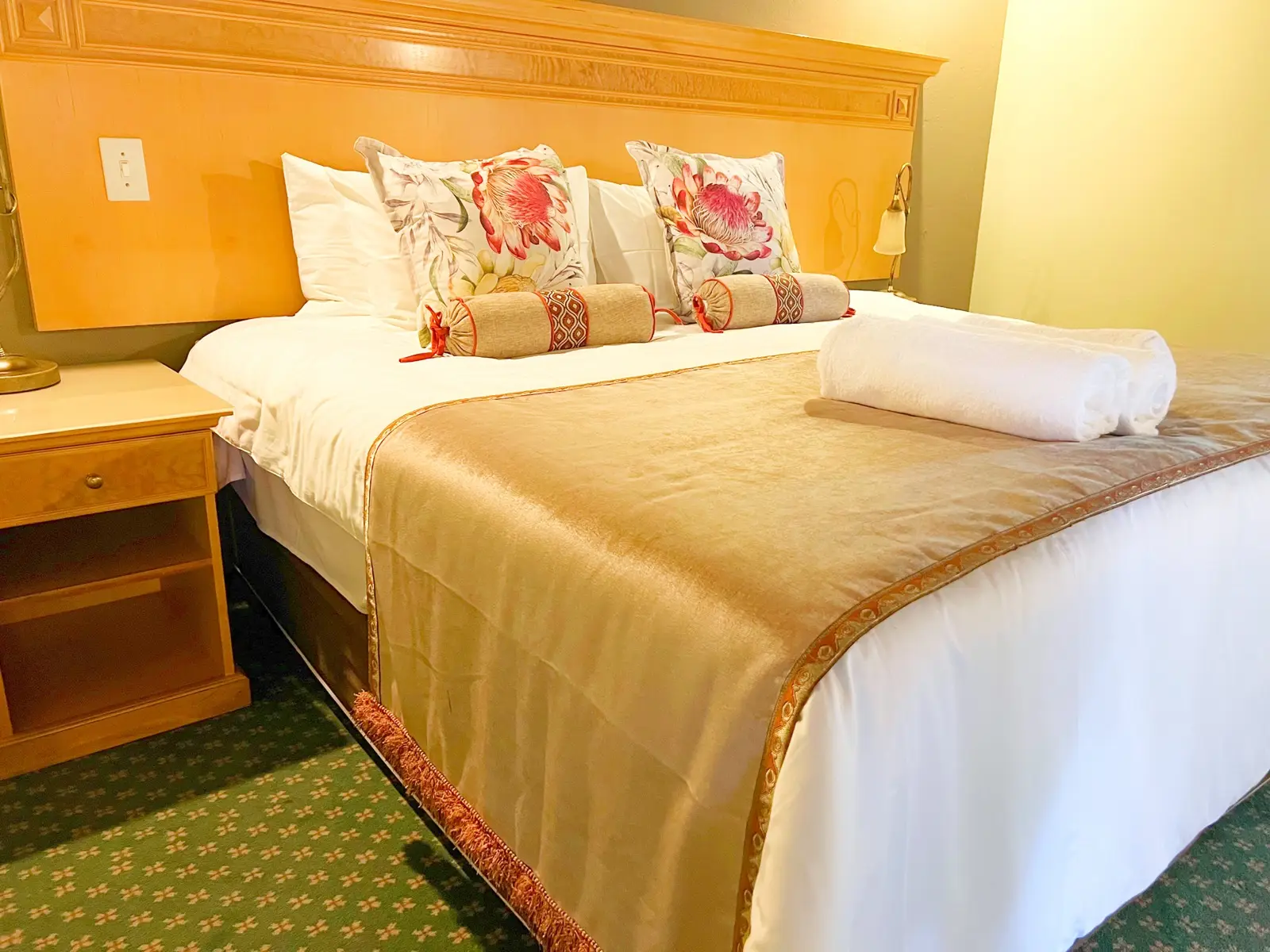 A neatly made bedroom with a gold throw and floral cushions.