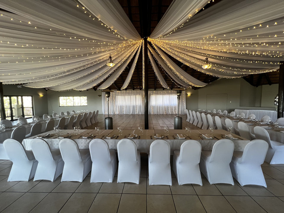A decorated thatched reception hall arranged with long tables and warm string lights.